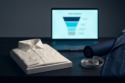 Folded off-the-rack shirt next to a tailored jacket with chalk marks and a measuring tape, a sales pipeline on a laptop behind