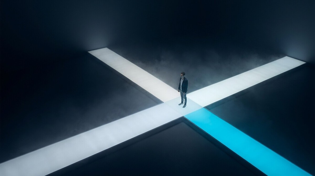 Lone person standing where illuminated walkways cross in a dark foggy space, one path glowing cyan