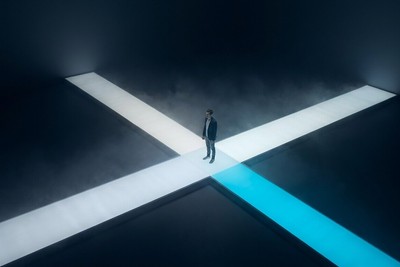 Lone person standing where illuminated walkways cross in a dark foggy space, one path glowing cyan
