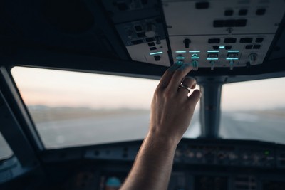 A pilot's hand flipping cockpit overhead switches lit in cyan before takeoff, with a runway visible through the windshield