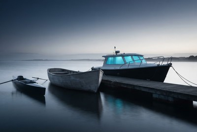 Three boats moored at a jetty in a misty harbor: a slim rowing skiff, a plain unfinished workboat and a motor launch with glowing cyan cabin windows