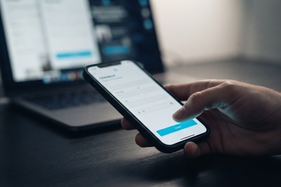 A thumb about to tap the cyan button on a phone checkout screen, with a laptop blurred in the background