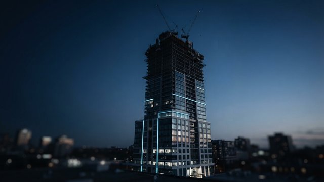 A skyscraper under construction at dusk with cranes on top and cyan light strips running up its lower floors