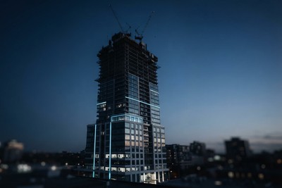 A skyscraper under construction at dusk with cranes on top and cyan light strips running up its lower floors