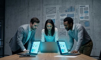 Three-person founding team leaning over laptops in front of a wall of wireframe sketches