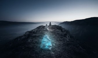 Ein leuchtender cyanfarbener Steinmann auf einem nebligen Bergpfad im Morgengrauen, zwei schlichte Steinmänner markieren den weiteren Weg