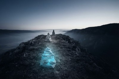 A glowing cyan stone cairn on a misty mountain ridge trail at dawn, with two plain cairns marking the path ahead