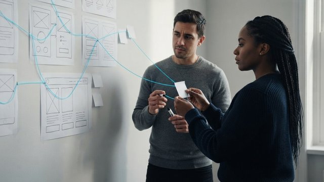 Two colleagues at a wall of wireframe sketches joined by a glowing cyan thread that traces a user journey, one holding a blank sticky note