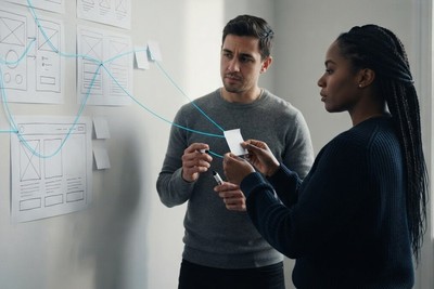 Two colleagues at a wall of wireframe sketches joined by a glowing cyan thread that traces a user journey, one holding a blank sticky note