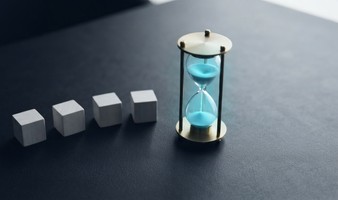 Hourglass on a desk beside four small labeled blocks representing the variables that set a project's pace